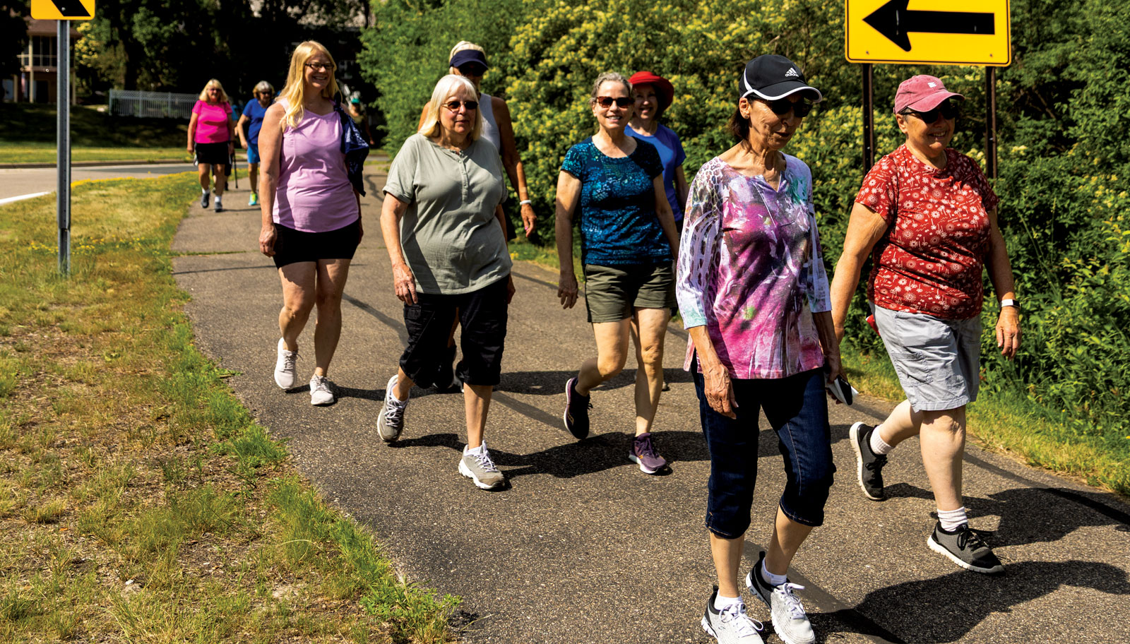 Members of the Bloomington Walking Club walk down an asphalt trail, with bushes and a grass boulevard on either side.